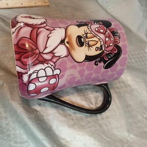 Puede incluir: Taza de cer&aacute;mica con Minnie Mouse vestida con un atuendo rosa con lunares blancos. La taza tiene un asa negra y un fondo morado claro con un patr&oacute;n de coraz&oacute;n. El dise&ntilde;o es una ilustraci&oacute;n de dibujos animados.