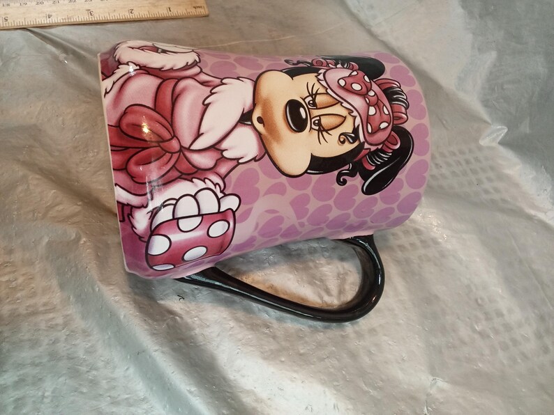 Puede incluir: Taza de cer&aacute;mica con Minnie Mouse vestida con un atuendo rosa con lunares blancos. La taza tiene un asa negra y un fondo morado claro con un patr&oacute;n de coraz&oacute;n. El dise&ntilde;o es una ilustraci&oacute;n de dibujos animados.