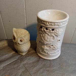 May include: A cream-colored owl figurine with golden eyes sits next to a white ceramic candle holder. The candle holder has a floral design and cut-out holes. A wooden ruler with inch markings is in the foreground.
