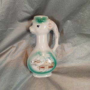 Könnte beinhalten: Dekorative weiße Keramikkrug mit hirschähnlichem Kopf und Griff. Der Krug hat einen grünen Rand und Boden, mit einer gemalten Landschaftsszene in Grün- und Rottönen. Der Krug ist etwa 15 cm hoch.
