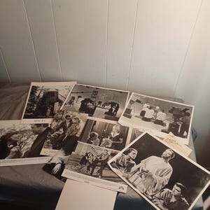 Peut inclure: Une collection de photographies vintage en noir et blanc, probablement issues d'une production cinématographique ou télévisuelle. Les images représentent diverses scènes, notamment des acteurs en costume et des prises de vue en coulisses. Les photos sont disposées sur une surface sombre.