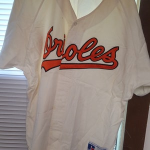 May include: White baseball jersey with orange "Orioles" lettering on the front. The jersey has a button-up front and a Russell Athletic logo on the bottom right.
