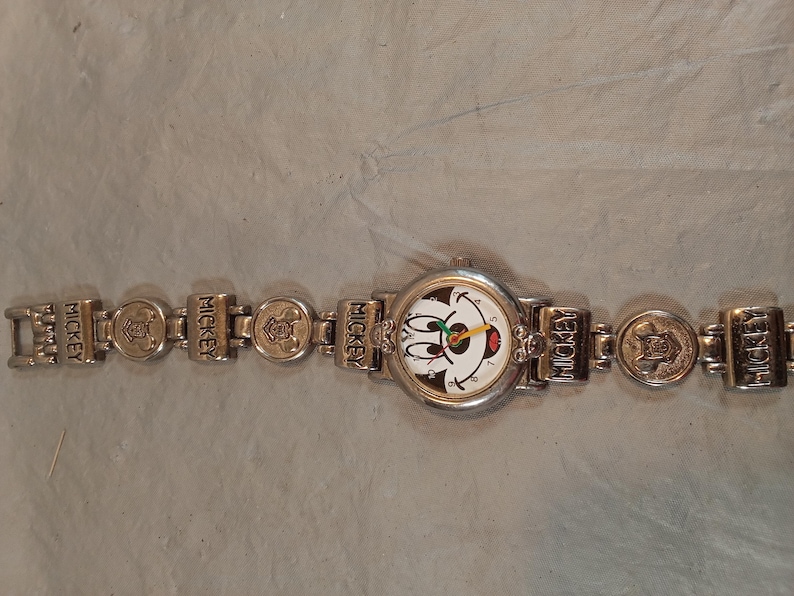 May include: A silver-toned analogue wristwatch featuring a round face with a Mickey Mouse graphic. The watch band consists of alternating rectangular links embossed with the word MICKEY and circular links featuring a raised character emblem.