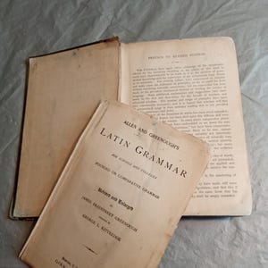 May include: An open vintage book with a tan cover titled "Latin Grammar" by Allen and Greenough, with the text "Revised and Enlarged" and "Founded on Comparative Grammar". The book is open to a page with text.