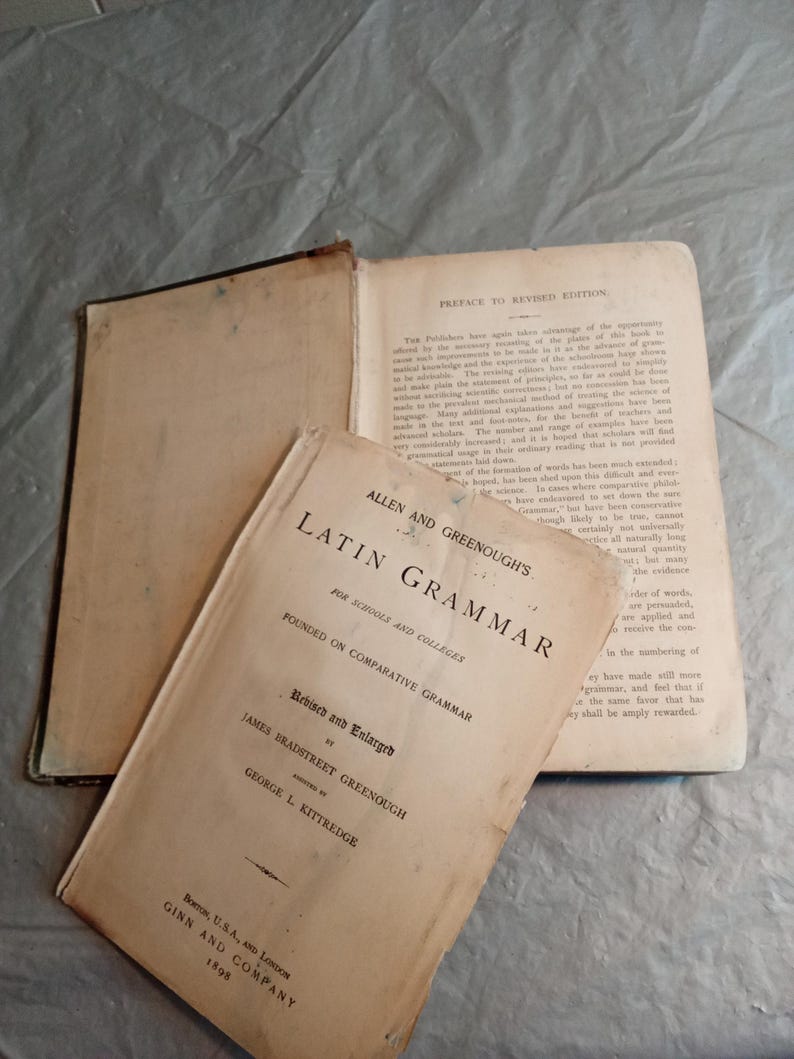 May include: An open vintage book with a tan cover titled "Latin Grammar" by Allen and Greenough, with the text "Revised and Enlarged" and "Founded on Comparative Grammar". The book is open to a page with text.