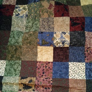 May include: A patchwork quilt featuring a mix of patterned squares in green, blue, brown, and burgundy. The quilt showcases floral, leaf, and geometric patterns, giving a rustic and cosy feel. The quilt is likely handmade.