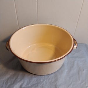 Puede incluir: Un tazón grande de esmalte blanco con un borde marrón y dos asas. El tazón está vacío y listo para ser usado.