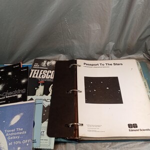 Peut inclure: Un classeur ouvert avec des livrets sur le thème de l'astronomie. Le classeur est ouvert sur une page intitulée "Passport To The Stars" avec une carte des étoiles. D'autres livrets incluent "Checking Around Telescope" et "Travel The Andromeda Galaxy at 10% OFF!"