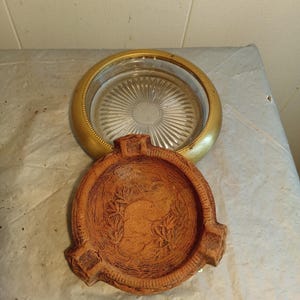 Peut inclure: Un cendrier et un dessous de verre sont présentés. Le cendrier est en terre cuite avec un motif de feuilles détaillé. Le dessous de verre a un bord doré et un centre en verre clair et strié. Les articles sont sur une surface claire.