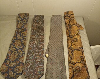 May include: Four neckties with various patterns are displayed. The ties include paisley and dotted designs in shades of blue, brown, and gold. The ties are laid out on a light-colored surface.