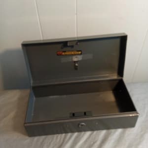 Peut inclure: Coffre-fort en métal gris ouvert, révélant un intérieur vide. Il possède une serrure et une clé, ainsi qu'un couvercle à charnière. La boîte est rectangulaire et repose sur une surface blanche.