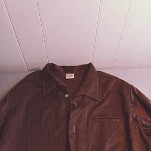 Op de afbeelding: Een bruin, lang mouwig overhemd met knopen en een klein zakje aan de linkerkant. Het overhemd heeft een subtiel herringbone-patroon en een wit label met de tekst "SUPERIOR QUALITY" erop.