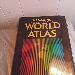 May include: A black hardcover book with gold lettering that reads "Hammond Ambassador World Atlas". The cover features a colorful world map with a rainbow striped design.