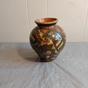 Puede incluir: Un jarrón de cerámica marrón con un patrón floral en tonos de amarillo, verde y marrón. El jarrón tiene un cuerpo redondeado y un cuello estrecho.