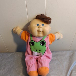 Könnte beinhalten: Eine Cabbage Patch Kid Puppe mit braunen Haaren und blauen Augen, die ein rosa-oranges Outfit mit einem grünen Frosch-Design trägt.
