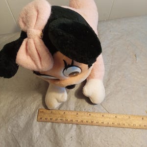 May include: A plush toy featuring a cartoon character with a pink body, black ears, and a pink bow. The toy has white paws and a smiling face. A wooden ruler is placed near the toy, measuring up to 30 inches.