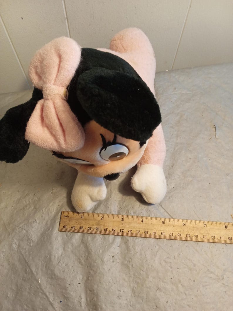 May include: A plush toy featuring a cartoon character with a pink body, black ears, and a pink bow. The toy has white paws and a smiling face. A wooden ruler is placed near the toy, measuring up to 30 inches.