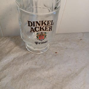 May include: A clear glass beer mug with the words "DINKEL ACKER" in black lettering. Below the text is a crest with red, green, and gold accents. The word "Privat" is printed below the crest. The mug has a handle and a thick base.