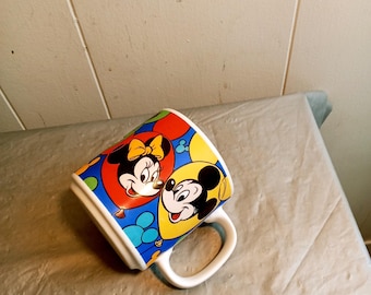 May include: A white ceramic mug featuring Mickey and Minnie Mouse. The mug has a colorful design with red, yellow, and blue accents. Mickey and Minnie are depicted in a heart-shaped design. The mug has a white handle.