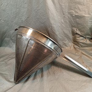Puede incluir: Un colador cónico de acero inoxidable con un cuerpo de malla fina, un borde ancho reforzado y un mango largo y plano que se extiende desde el lateral. La superficie metálica tiene un acabado pulido con perforaciones visibles en el cono.