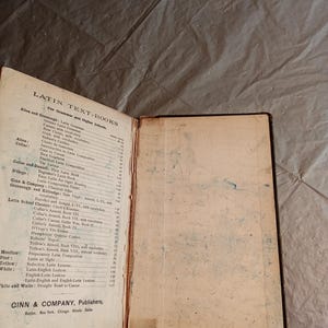 May include: An open vintage Latin textbook with a worn cover and pages. The left page displays a detailed table of contents for grammar and higher schools. The book is published by Ginn & Company.