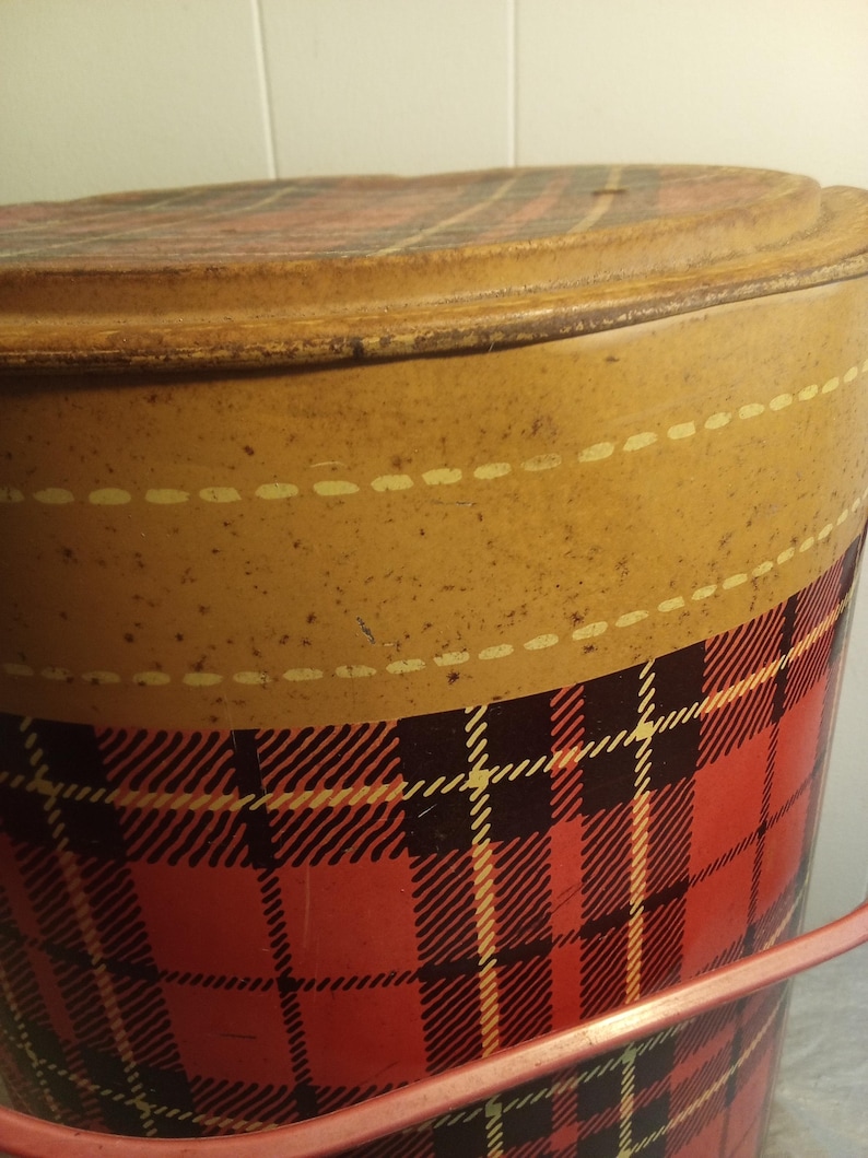 May include: Vintage metal bucket with a lid. The bucket features a red and black plaid pattern on the lower portion and a tan upper section with a dotted line design. The lid also has a plaid pattern. A pink handle is visible.