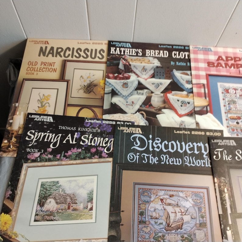 Pode incluir: Uma cole&ccedil;&atilde;o de oito livros de padr&otilde;es de ponto cruz com capas coloridas. Os t&iacute;tulos incluem "Narcissus Old Print Collection Book 1", "Kathie's Bread Cloths by Kathie R", "Spring At Stoneg Book 3", "Discovery of the New World" e "Apple Sampler".
