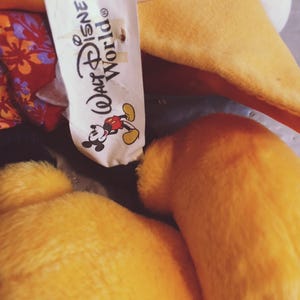 Puede incluir: Primer plano de un peluche con pelaje amarillo y una etiqueta blanca. La etiqueta dice "Walt Disney World" con un gr&aacute;fico de Mickey Mouse. El juguete tiene un aspecto suave y texturizado.
