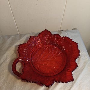 May include: A red glass serving dish in the shape of a leaf. The dish has a handle and detailed veining throughout. The edges are scalloped. The dish is likely vintage and is a vibrant shade of red.