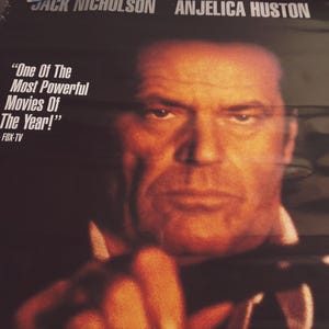 May include: A movie poster featuring Jack Nicholson and Anjelica Huston. The poster has a black background with white text that reads "One Of The Most Powerful Movies Of The Year!" and "FOX-TV". Jack Nicholson is pictured in the foreground, looking directly at the viewer with a serious expression. He is wearing a dark jacket and has a hand near his face.