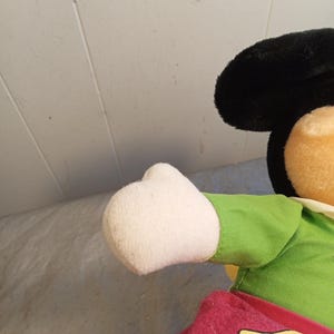 May include: A plush Mickey Mouse doll, featuring a black head with large ears, a tan face, and a white gloved hand. The doll wears a green shirt and red shorts with yellow accents. The background is a light gray surface.