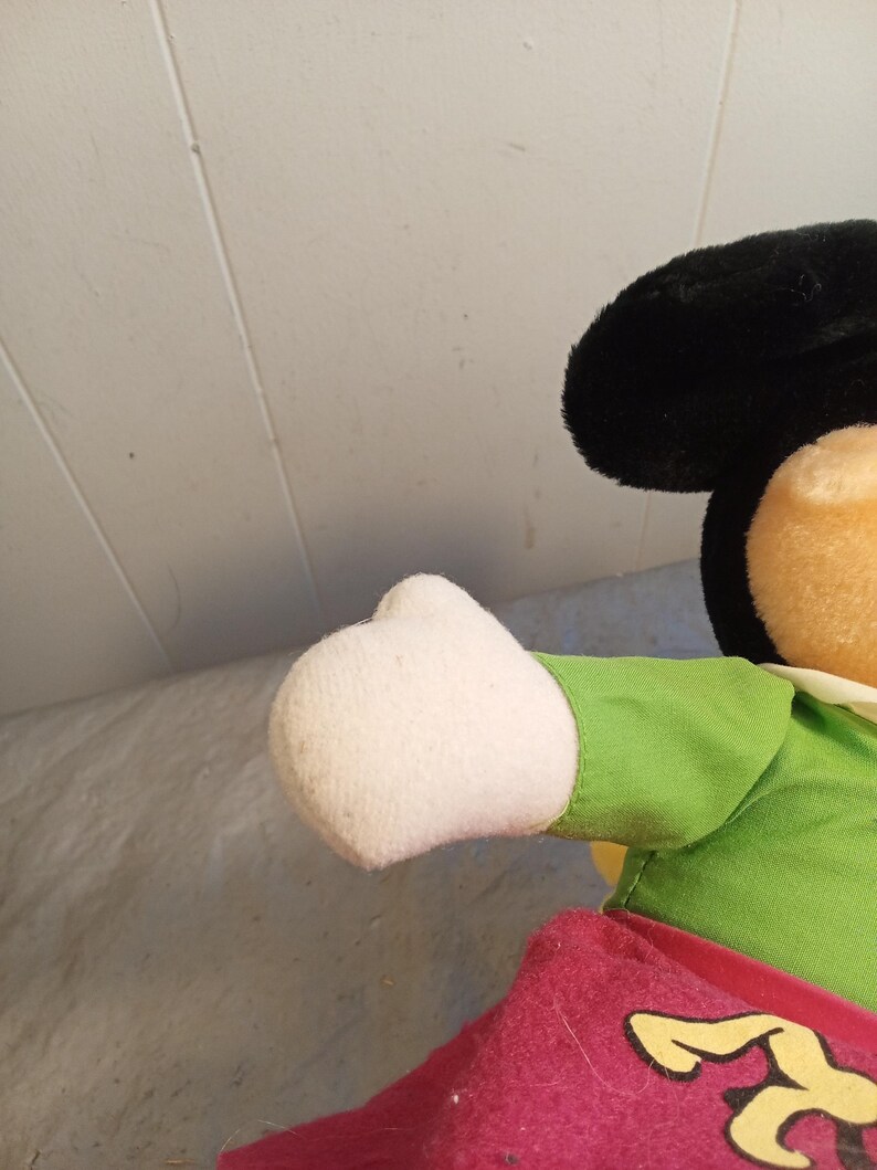 May include: A plush Mickey Mouse doll, featuring a black head with large ears, a tan face, and a white gloved hand. The doll wears a green shirt and red shorts with yellow accents. The background is a light gray surface.