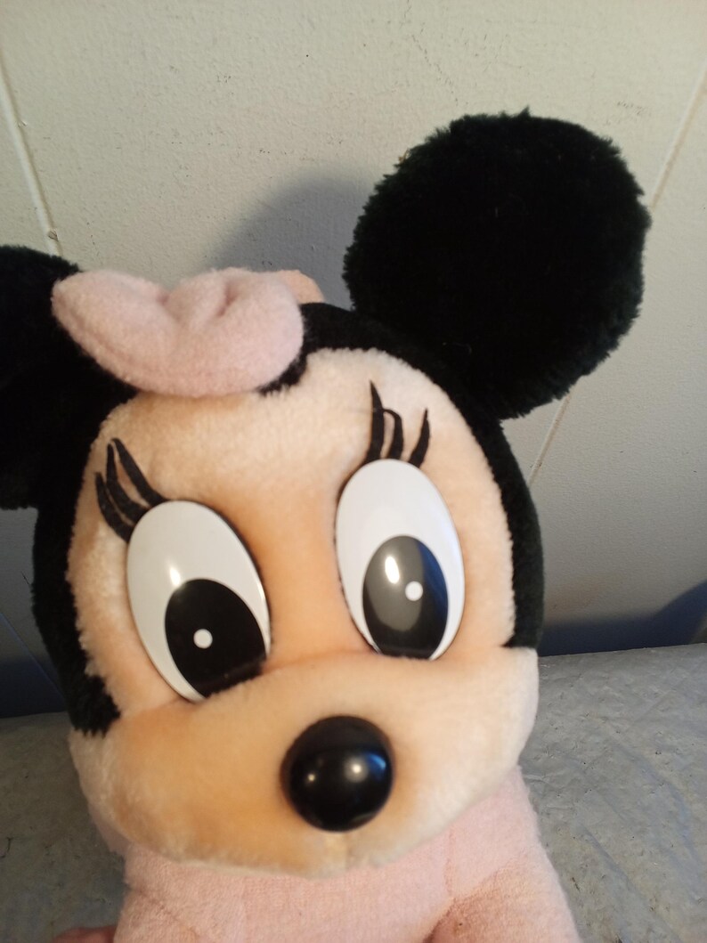 May include: A plush Minnie Mouse doll with a pink bow, black ears, and large, expressive eyes. The doll has a pink face and body, with a black nose and eyelashes. A classic Disney character.