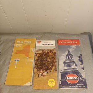 May include: Three vintage travel maps: New York City, Nebraska, and Philadelphia. The New York map is yellow with a state outline. The Nebraska map features a person on a horse. The Philadelphia map shows a clock tower with an AMOCO logo.
