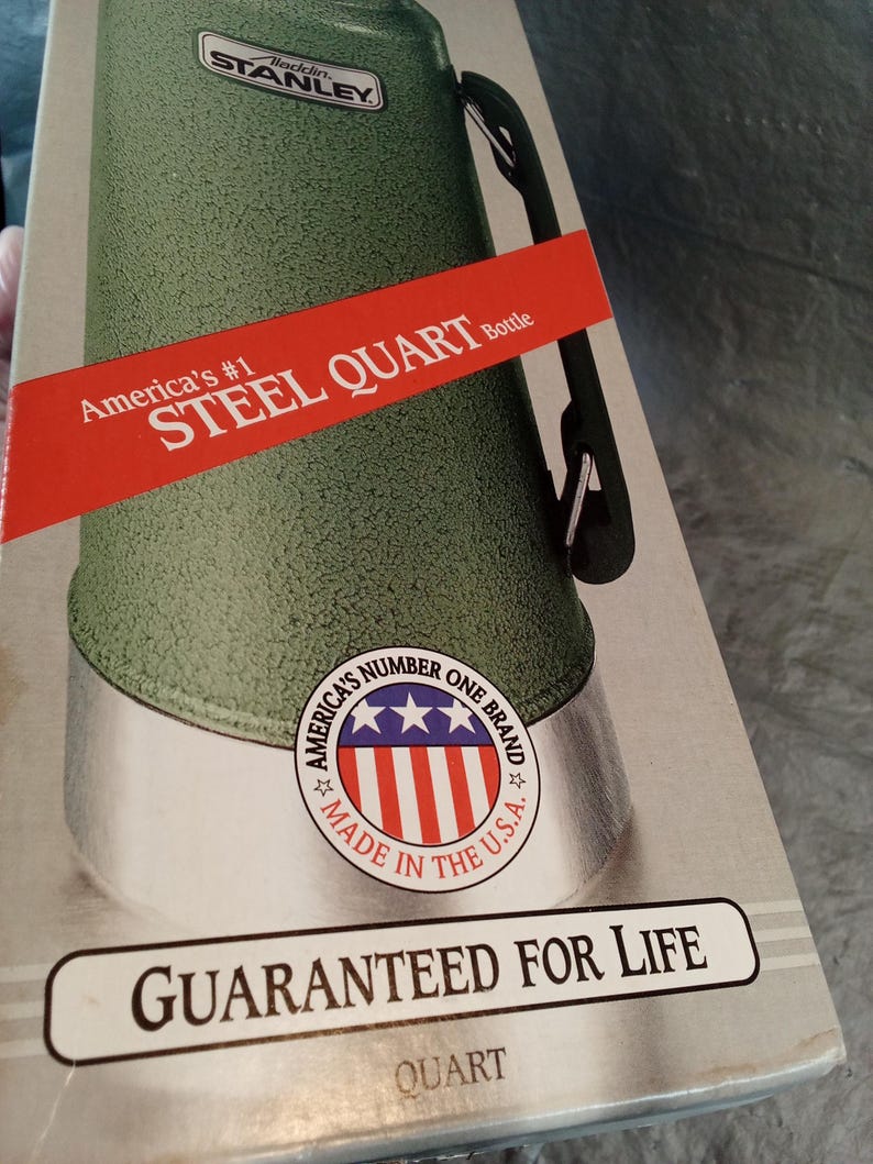 May include: A green Stanley Aladdin steel quart bottle in its original packaging. The box features a red banner with the text America's #1 STEEL QUART Bottle, a circular logo with stars and stripes, and the text GUARANTEED FOR LIFE at the bottom.