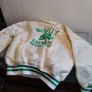 Peut inclure: Une veste bomber en satin blanc avec des poignets et un col rayés vert et blanc. Le dos de la veste présente un logo de trèfle vert avec le texte "McGill's Express Paterson, N.J."