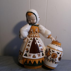 May include: A ceramic figurine of a girl in a white dress with a brown and yellow skirt and a green and yellow hat. She is holding a brown and yellow ceramic pot with a bird design.