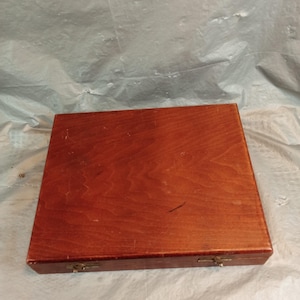 May include: A rectangular wooden storage box with a smooth, reddish-brown finish and visible wood grain. The box features two small, metallic latches on the front edge for securing the lid, showing signs of light wear on the surface.