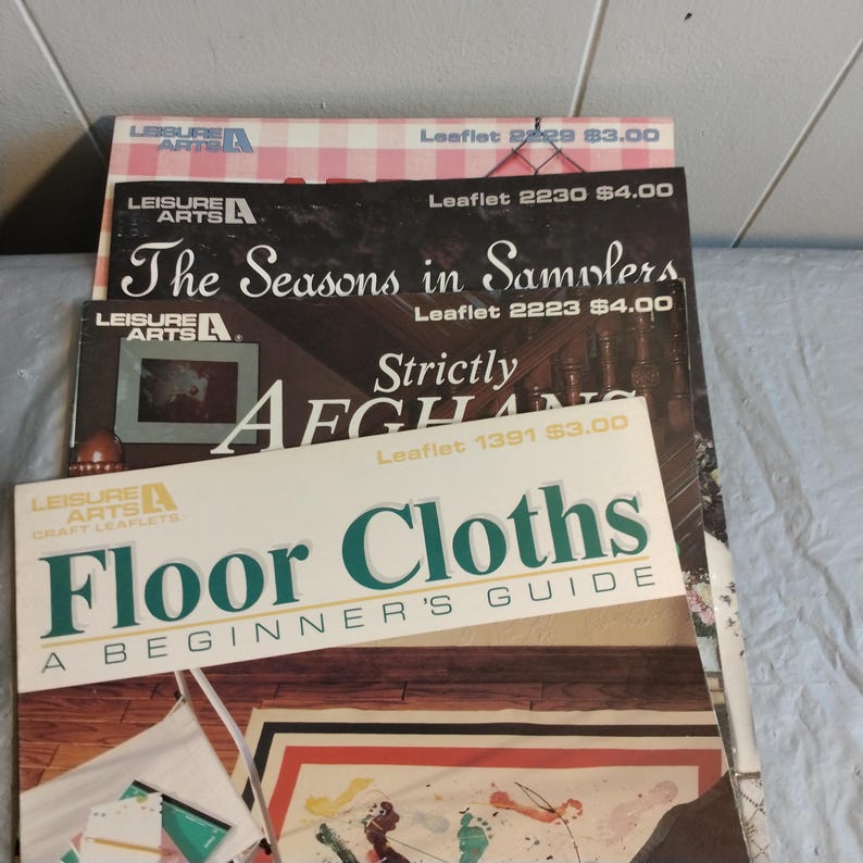 Pode incluir: Uma pilha de cinco folhetos de artesanato da Leisure Arts. O folheto superior &eacute; intitulado "Floor Cloths: A Beginner's Guide". Os outros folhetos s&atilde;o intitulados "The Seasons in Samplers", "Strictly Afghans", e "Leaflet 2229" e "Leaflet 2230".