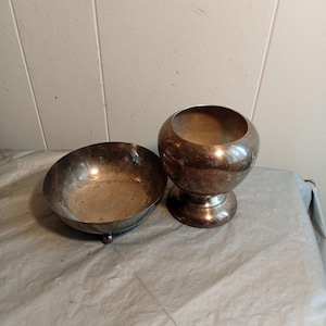 Puede incluir: Dos cuencos antiguos de color plateado. Uno es un cuenco poco profundo con forma redondeada y un pie pequeño. El otro es un cuenco con pedestal con la parte superior redonda y una base acampanada. Ambos cuencos tienen un aspecto deslustrado.