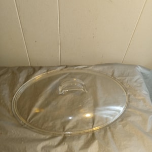 Puede incluir: Una tapa de vidrio transparente de forma ovalada con un pequeño asa. La tapa está diseñada para una olla o sartén. El material transparente permite ver el contenido. La tapa está sobre una superficie arrugada de color claro.