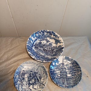 Könnte beinhalten: Drei blau-weiße Keramikplatten mit historischen Szenen. Jede Platte zeigt ein anderes Design: eine Pferdekutsche, Reiter auf Pferden und ein Gebäude mit hohem Turm. Die Teller haben eine klassische, Vintage-Ästhetik.