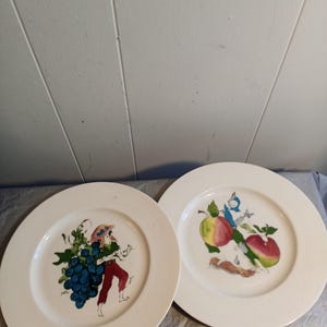 May include: Two white ceramic plates with colorful illustrations. One plate features a figure carrying grapes, the other has apples and a small figure. The plates have a wide rim.