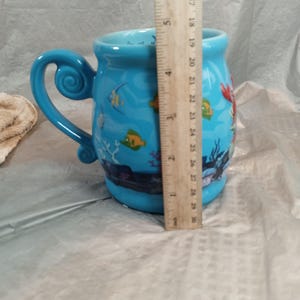 May include: A blue ceramic mug with a whimsical handle and a sea-themed design. The mug features illustrations of fish, coral, and a mermaid. The mug measures approximately 4 inches in diameter and 4 inches in height.