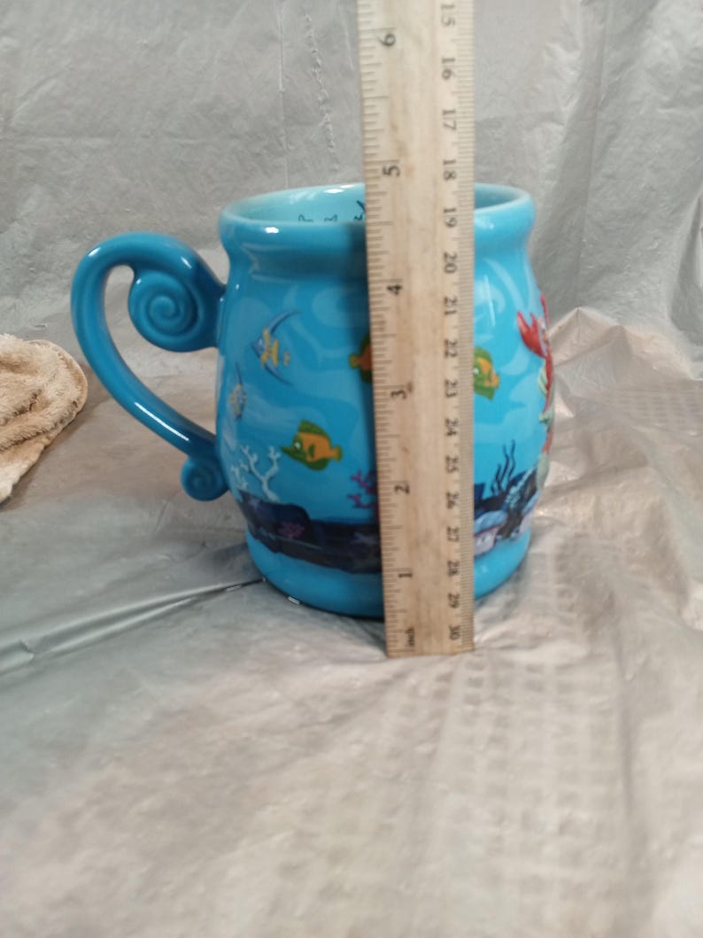 May include: A blue ceramic mug with a whimsical handle and a sea-themed design. The mug features illustrations of fish, coral, and a mermaid. The mug measures approximately 4 inches in diameter and 4 inches in height.