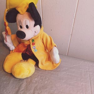 Puede incluir: Un mu&ntilde;eco de peluche de Mickey Mouse con birrete y toga amarillos. El mu&ntilde;eco tiene orejas negras, cara blanca y nariz negra. La toga tiene bordada la palabra "2000". La borla del gorro es roja y dorada.