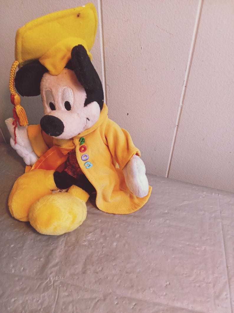 Puede incluir: Un mu&ntilde;eco de peluche de Mickey Mouse con birrete y toga amarillos. El mu&ntilde;eco tiene orejas negras, cara blanca y nariz negra. La toga tiene bordada la palabra "2000". La borla del gorro es roja y dorada.