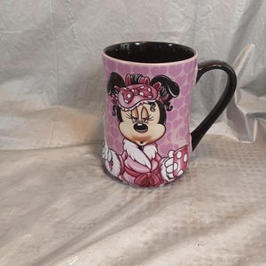 Puede incluir: Taza de cer&aacute;mica con Minnie Mouse con bata y gorro de dormir rosas. La taza tiene un asa e interior negros, con un fondo rosa cubierto de peque&ntilde;as formas de coraz&oacute;n. El dise&ntilde;o es una ilustraci&oacute;n de dibujos animados.
