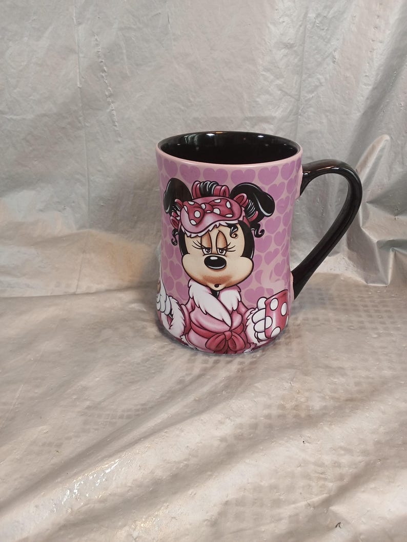 Puede incluir: Taza de cer&aacute;mica con Minnie Mouse con bata y gorro de dormir rosas. La taza tiene un asa e interior negros, con un fondo rosa cubierto de peque&ntilde;as formas de coraz&oacute;n. El dise&ntilde;o es una ilustraci&oacute;n de dibujos animados.