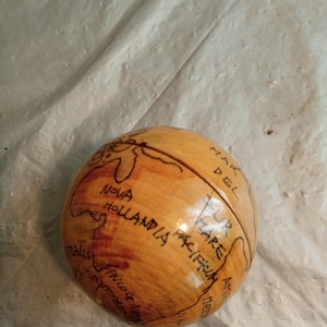 Puede incluir: Un pequeño globo terráqueo de madera con continentes y texto dibujados a mano. El globo es de color marrón claro con marcas y texto marrón más oscuro, incluyendo "Nova Hollandia" y "Mare Pacificum".